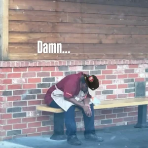A Popeyes employe wearing a Popeyes uniform and cap sits hunched over on a bench against a brick wall, conveying exhaustion because of extensively delivering Popeyes chicken sandwiches. Text above reads, “Damn...”